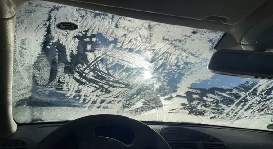 Vetro dell'auto coperto di brina, con segni di tergicristallo. Un sole pallido filtra attraverso il ghiaccio, rendendo difficile vedere. Ecco come sbrinare il vetro della macchina.