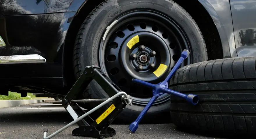 Kit per cambiare una ruota: cric, chiave a stella e ruota di scorta accanto a un'auto nera.