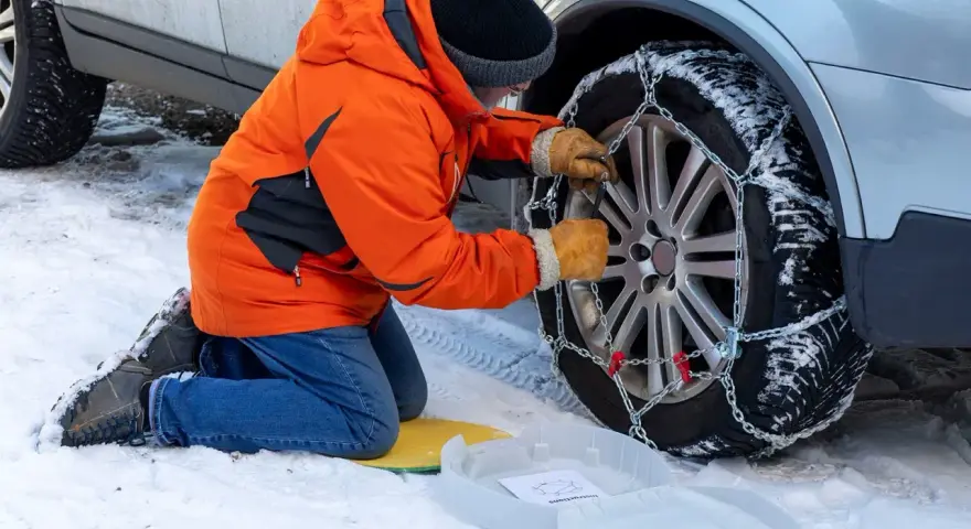 Uomo in giacca arancione monta catene da neve su ruota posteriore. Le catene da neve devono essere montate sulle ruote posteriori per la massima aderenza.