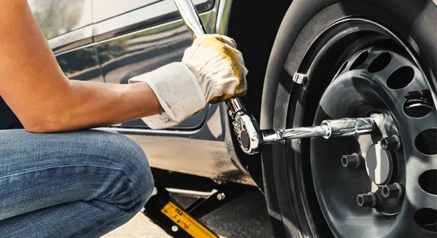 Persona con guanti gialli usa una chiave per serrare i bulloni della ruota di un'auto sollevata da un cric. Il significato di "nm" è la coppia di serraggio.