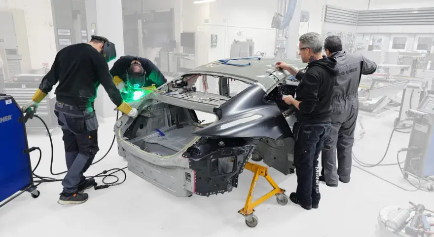 Operai saldano il telaio macchina in officina. Uno di loro usa una saldatrice, mentre gli altri osservano e lavorano sulla carrozzeria.