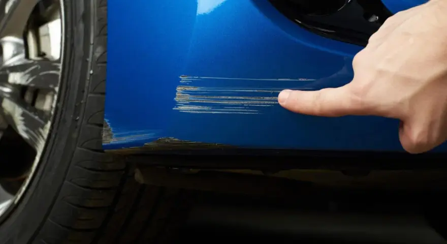 Dito, un dito indica profondi graffi sul parafango blu di un'auto, segno che necessita di riparazione vernice auto.