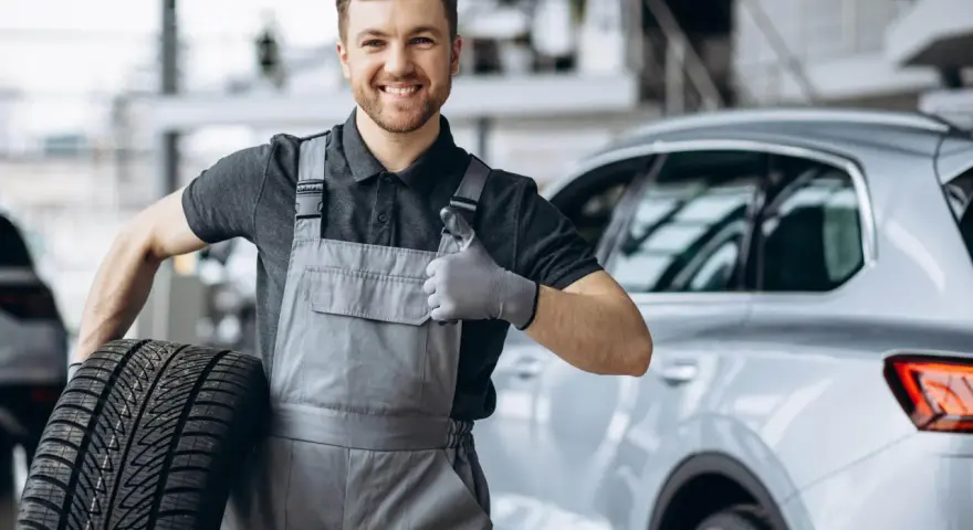 Meccanico sorridente con una gomma primo equipaggiamento in mano, fa il pollice in su accanto a un'auto.