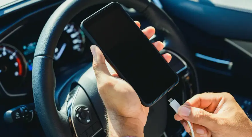 Mano che collega un cavo a uno smartphone, con il volante e il quadro strumenti dell'auto sullo sfondo. Se la batteria è scarica, il quadro si accende.