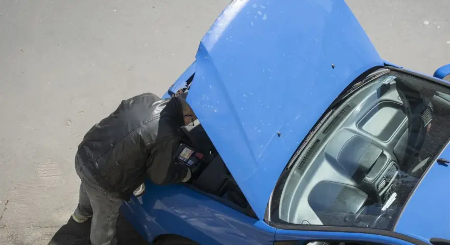 Uomo con giacca in pelle controlla la **macchina ferma** con il cofano aperto, forse un problema alla **batteria**.