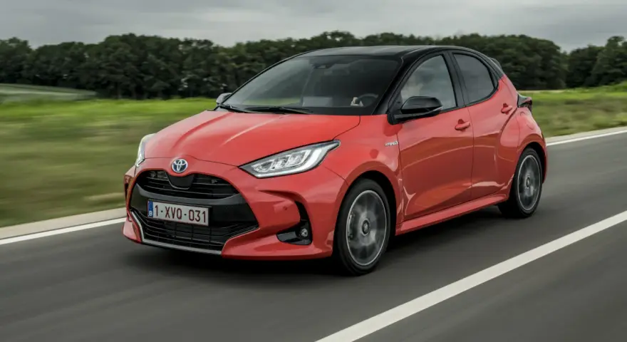 Auto ibrida rossa in movimento su strada, con sfondo sfocato di alberi e cielo nuvoloso. La scritta "Hybrid" è visibile sul fianco.