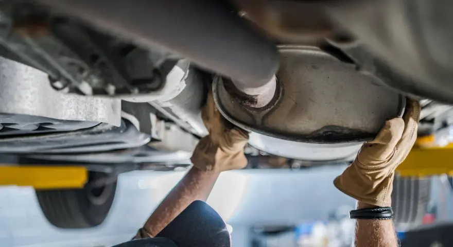 Mani guantate lavorano sul sistema di scarico di un'auto, forse sostituendo il catalizzatore auto.