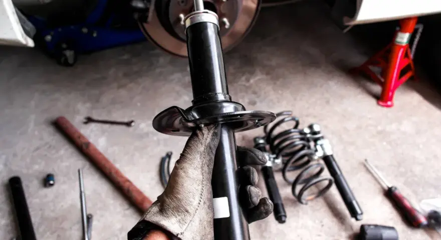 Gloved hand holding a car shock absorber with tools and springs on the ground.