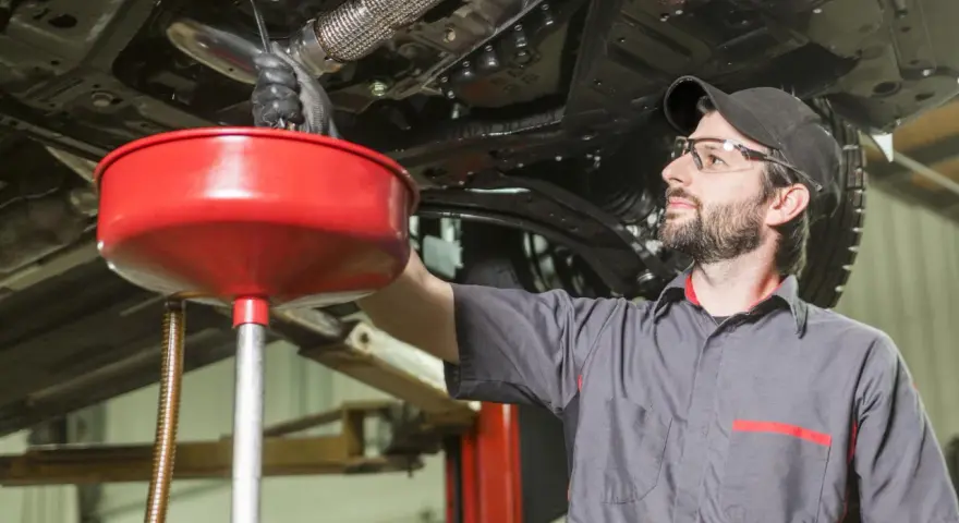 Meccanico con occhiali protettivi e cappellino lavora sotto un'auto sollevata, raccogliendo liquidi in un imbuto rosso. Valutazione pro e contro lavaggio interno motore.