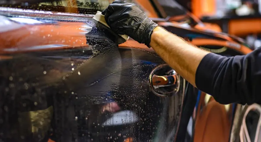 Mano con guanto nero pulisce la carrozzeria lucida di un'auto, rimuovendo la resina con un panno umido e schiumoso.