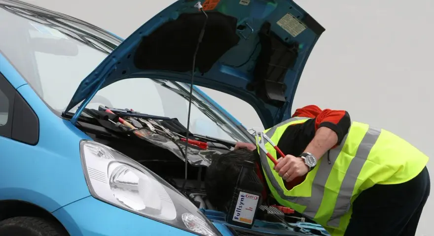 Meccanico con giubbotto ad alta visibilità lavora sul motore di un'auto blu. Nonostante il silenzio delle auto elettriche, il rumore degli attrezzi è forte.
