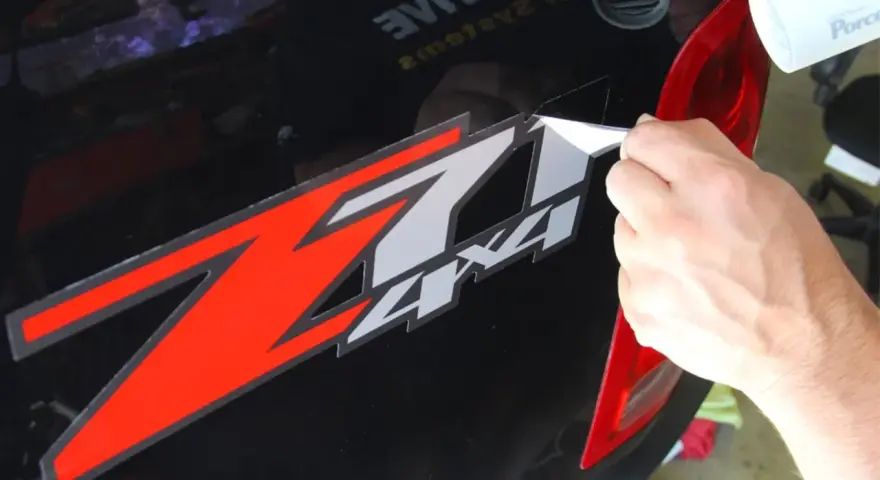Come togliere colla adesivi: una mano rimuove un adesivo rosso e bianco "Z 4x4" da un'auto nera, usando un phon.