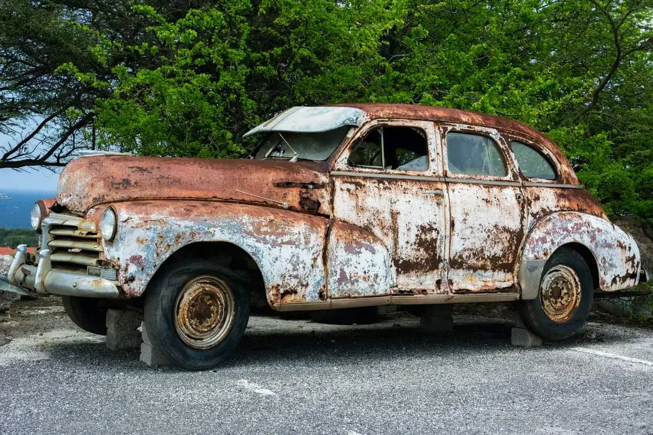 La scocca dell'auto, arrugginita e scrostata, giace abbandonata sotto alberi verdi.