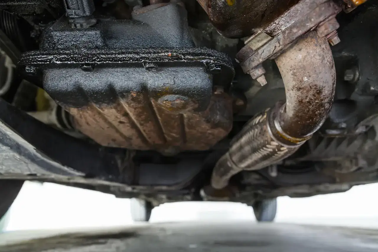 Sotto un'auto, il carter dell'olio è coperto di grasso e presenta segni di perdita olio motore. Si vedono anche parti del sistema di scarico.