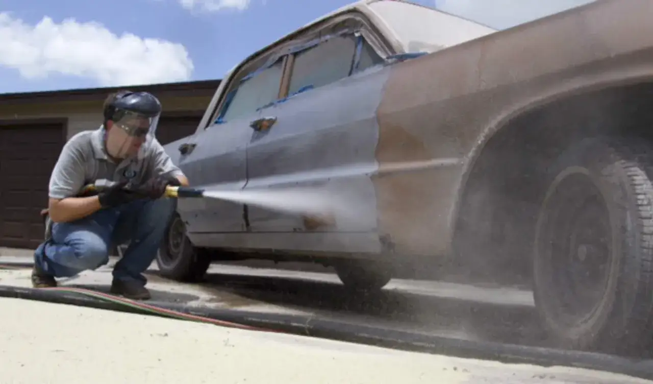 Uomo con maschera protettiva esegue soda blasting su un'auto d'epoca, rimuovendo ruggine e vernice.