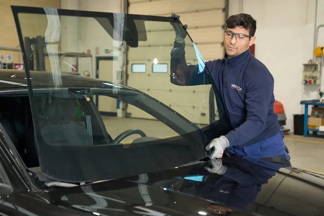 Tecnico con occhiali protettivi posiziona un parabrezza nuovo su un'auto. Si attende il tempo asciugatura colla parabrezza per completare l'installazione.