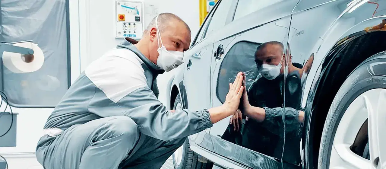 Un operaio con mascherina esegue la carteggiatura auto, controllando la finitura della carrozzeria lucida.