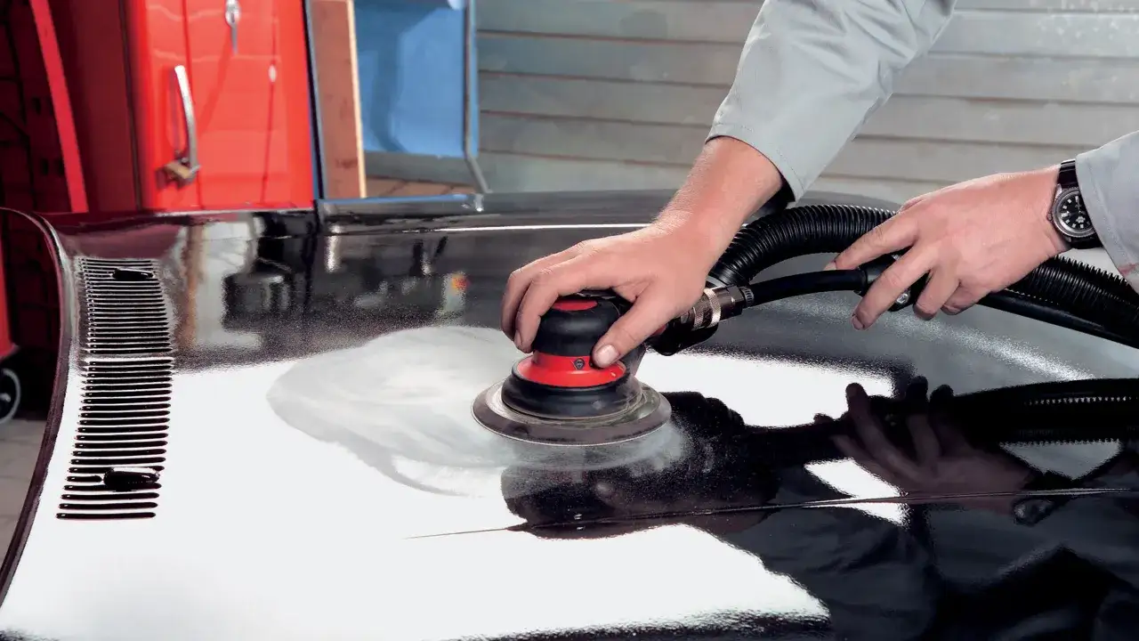 Mani esperte levigano una superficie lucida, forse un'auto, con una levigatrice. Lo stucco nero è quasi invisibile sotto la finitura perfetta.