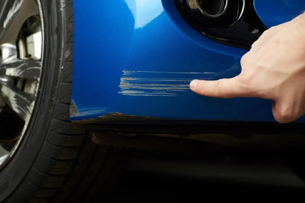 Dito, un dito indica profondi graffi sul parafango blu di un'auto, segno che necessita di riparazione vernice auto.