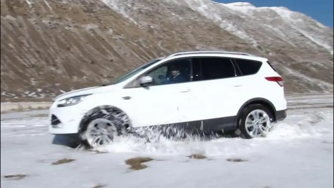 Bianca auto SUV affronta la neve con trazione posteriore, sollevando spruzzi. Montagne innevate sullo sfondo.