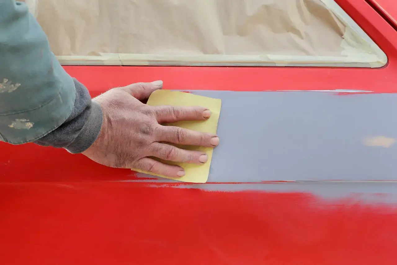 Mano che carteggia una zona grigia su un'auto rossa, preparando la superficie per la verniciatura. Questo &egrave; un passaggio fondamentale per capire cos'&egrave; il primer vernice.
