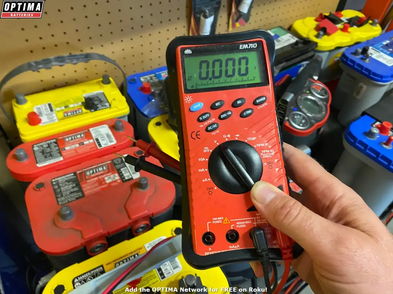 Misurazione del voltaggio di una batteria auto con un multimetro. Diverse batterie Optima sono visibili sullo sfondo.
