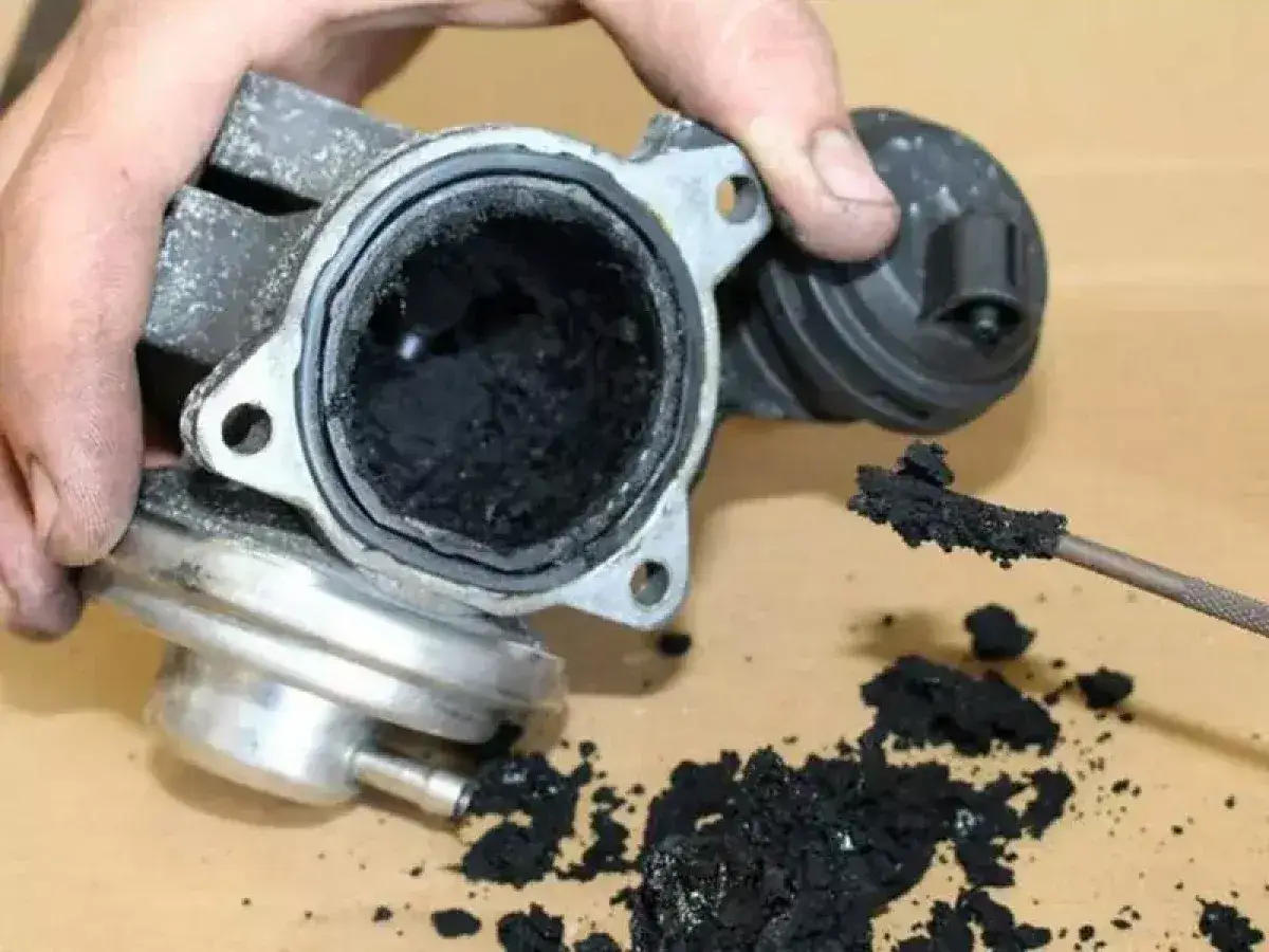 Mano che mostra una valvola EGR sporca, incrostata di fuliggine nera. Un attrezzo rimuove residui carboniosi.