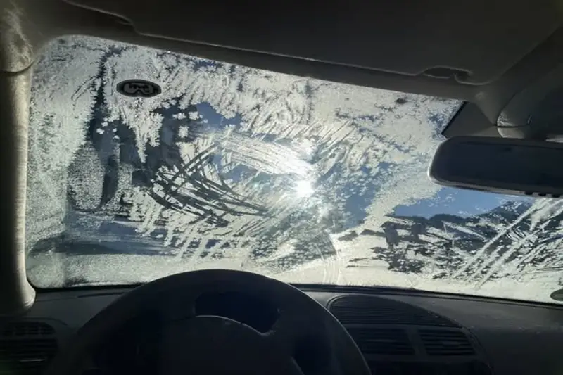Vetro dell'auto coperto di brina, con segni di tergicristallo. Un sole pallido filtra attraverso il ghiaccio, rendendo difficile vedere. Ecco come sbrinare il vetro della macchina.