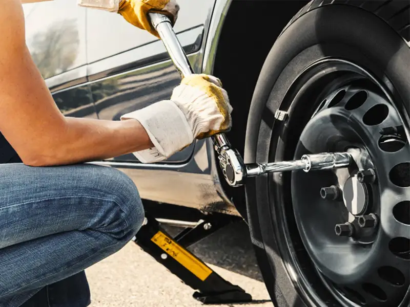 Persona con guanti gialli usa una chiave per serrare i bulloni della ruota di un'auto sollevata da un cric. Il significato di "nm" è la coppia di serraggio.